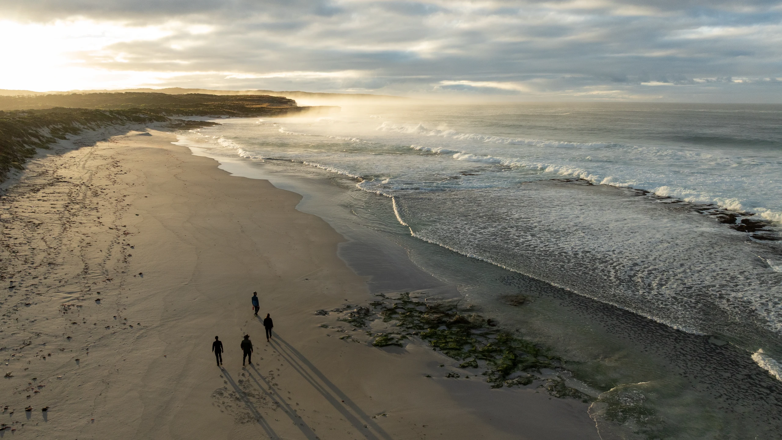 Four walkers and their long shadows on a broad beach, with sunlit cliffs, sea mist and breaking waves along the coastline.