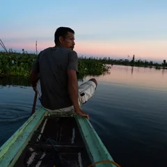 A man sitting at the bow of a narrow wooden boat on calm water at dusk.