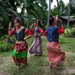 Three people in colorful beaded clothing dancing among palm and banana trees.