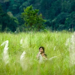 A woman standing among tall green grasses with forest behind her.