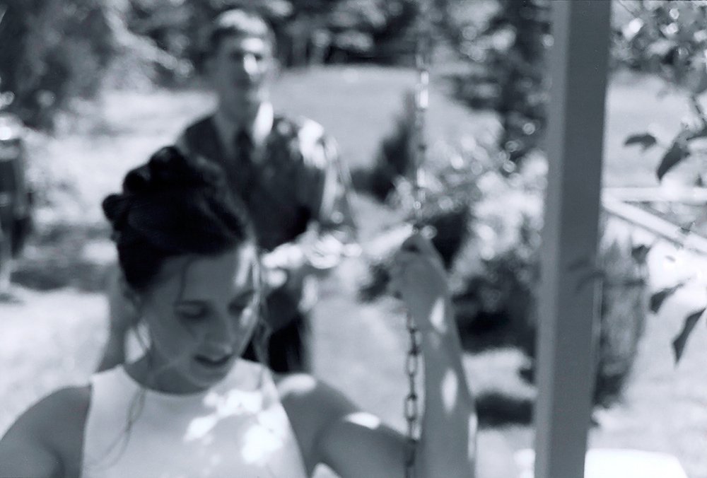 Black-and-white wedding photograph of a bride on a swing, with the groom behind her.